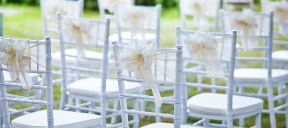 A close-up of white wedding chairs with white decorative linens attached to their backs.