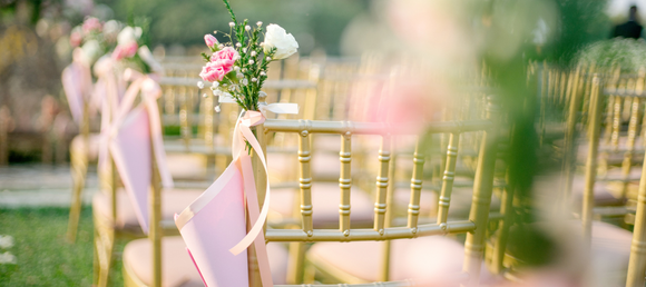 A close-up of gold wedding chairs with flowers and pink decorative linens attached to their backs.
